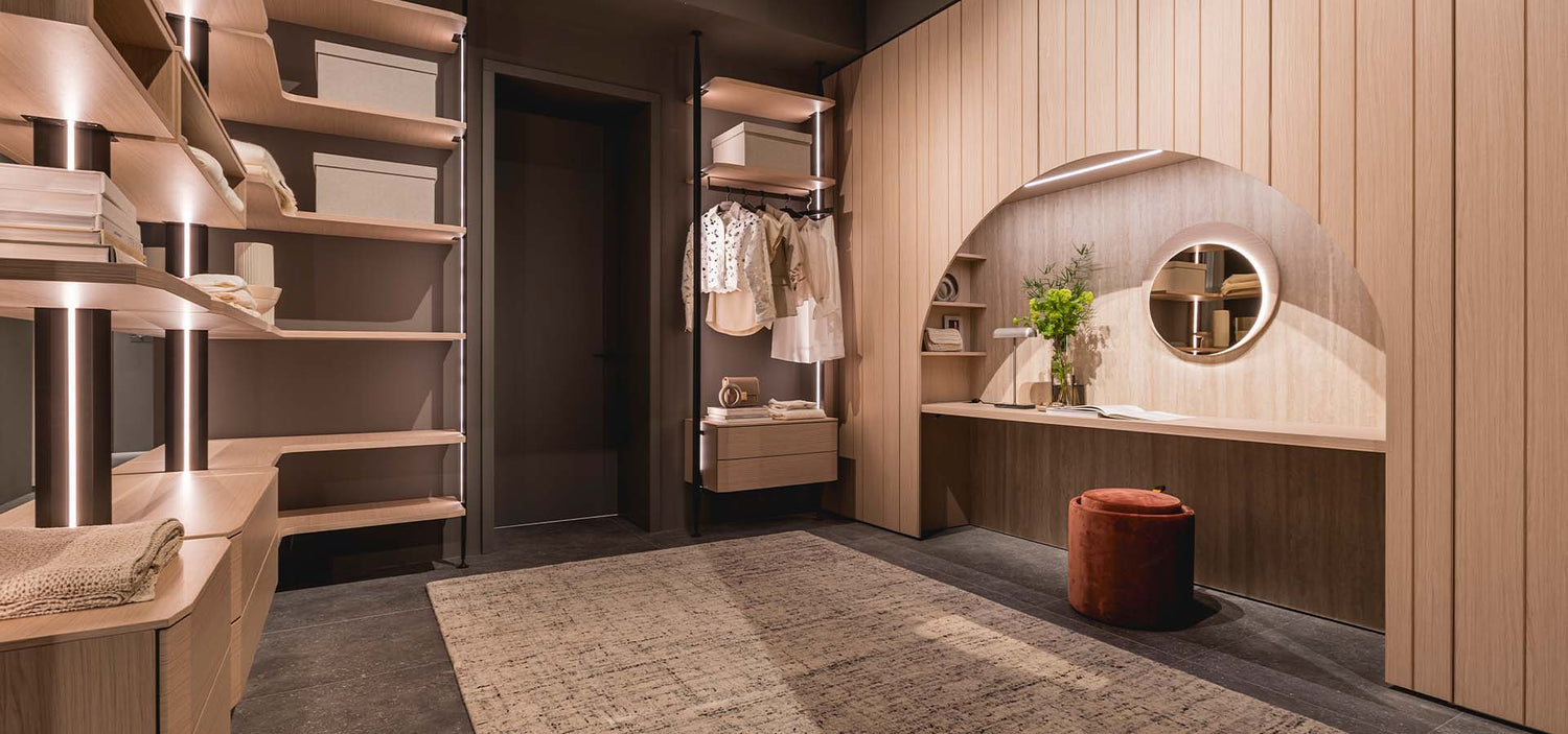 Cabina armadio di design con mensole in legno chiaro, zona trucco con specchio rotondo illuminato e pouf in velluto rosso, illuminazione LED integrata.