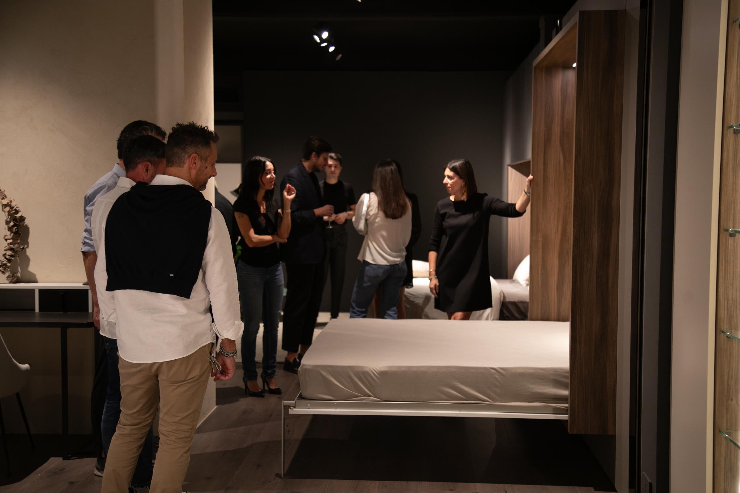 Persone durante un evento in showroom di arredamento, con una donna che illustra un letto a scomparsa in legno a un gruppo di visitatori