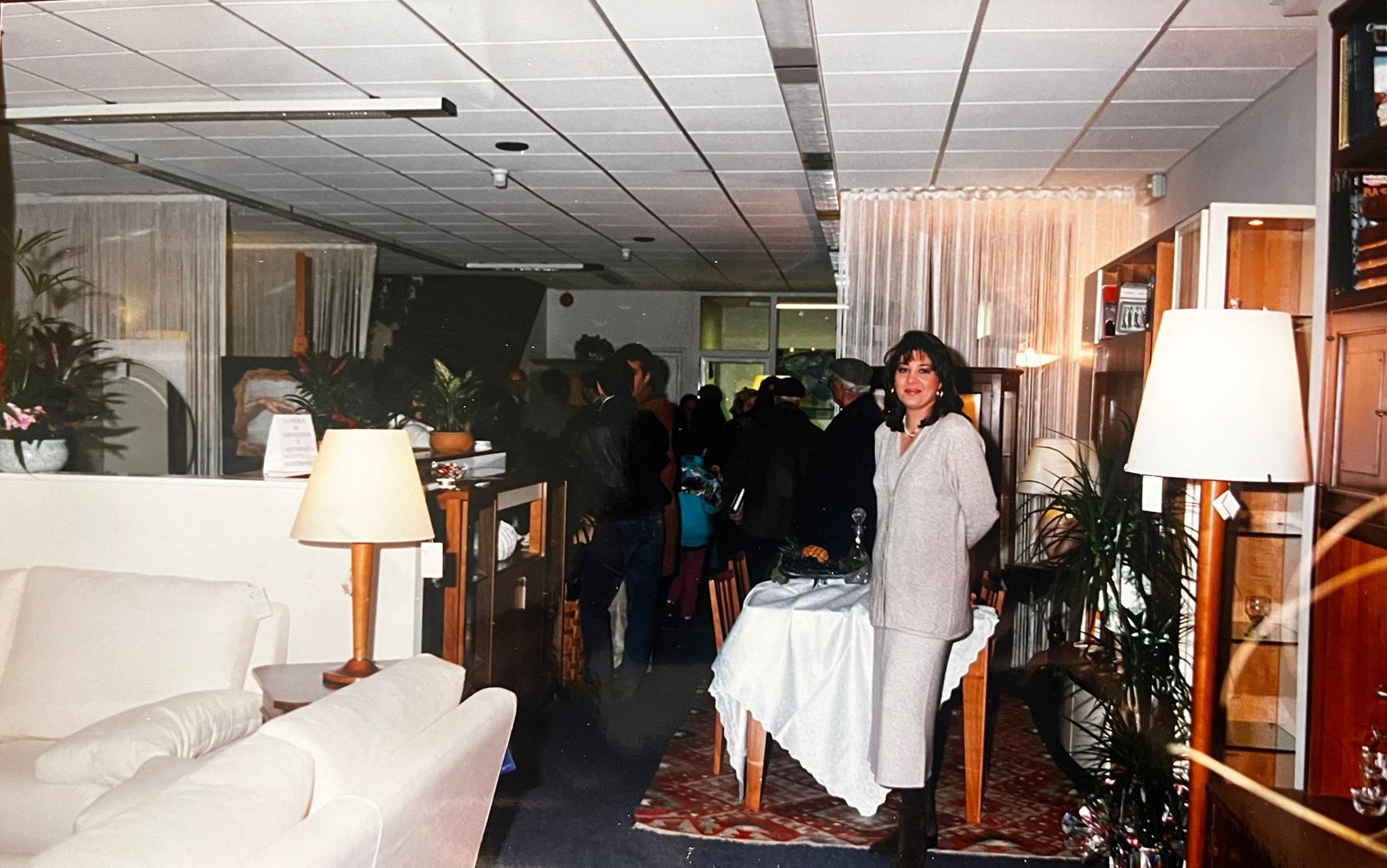 vento in negozio di mobili con clientela e arredamenti esposti, donna in abito chiaro accanto a tavolo apparecchiato e lampada da tavolo su tappeto decorativo.