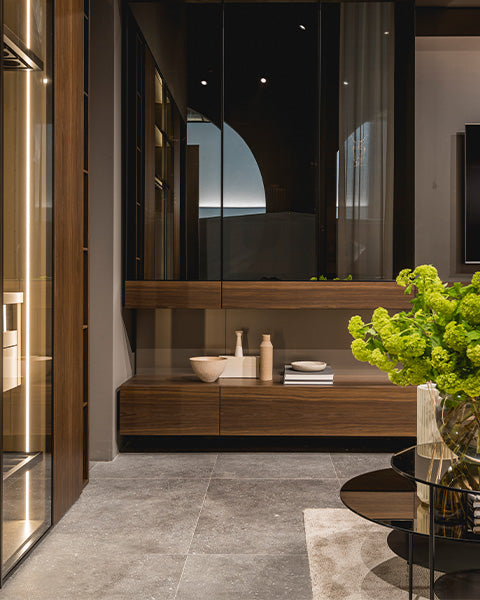 Soggiorno elegante con mobile basso in legno scuro e ante in vetro, tavolino in vetro nero con vaso di fiori verdi e pavimento in pietra grigia.