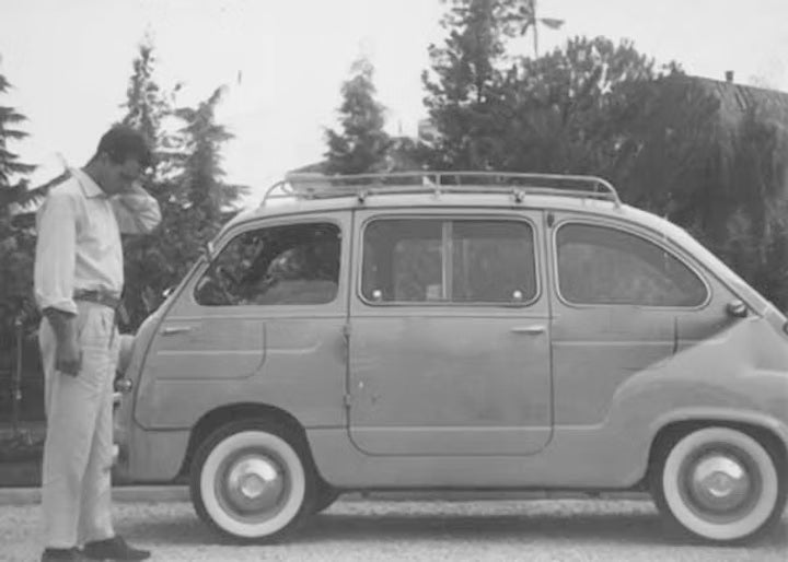 Foto storica in bianco e nero di un uomo accanto a una Fiat 600 Multipla, simbolo dell’Italia degli anni ‘60 e dei viaggi d’epoca.