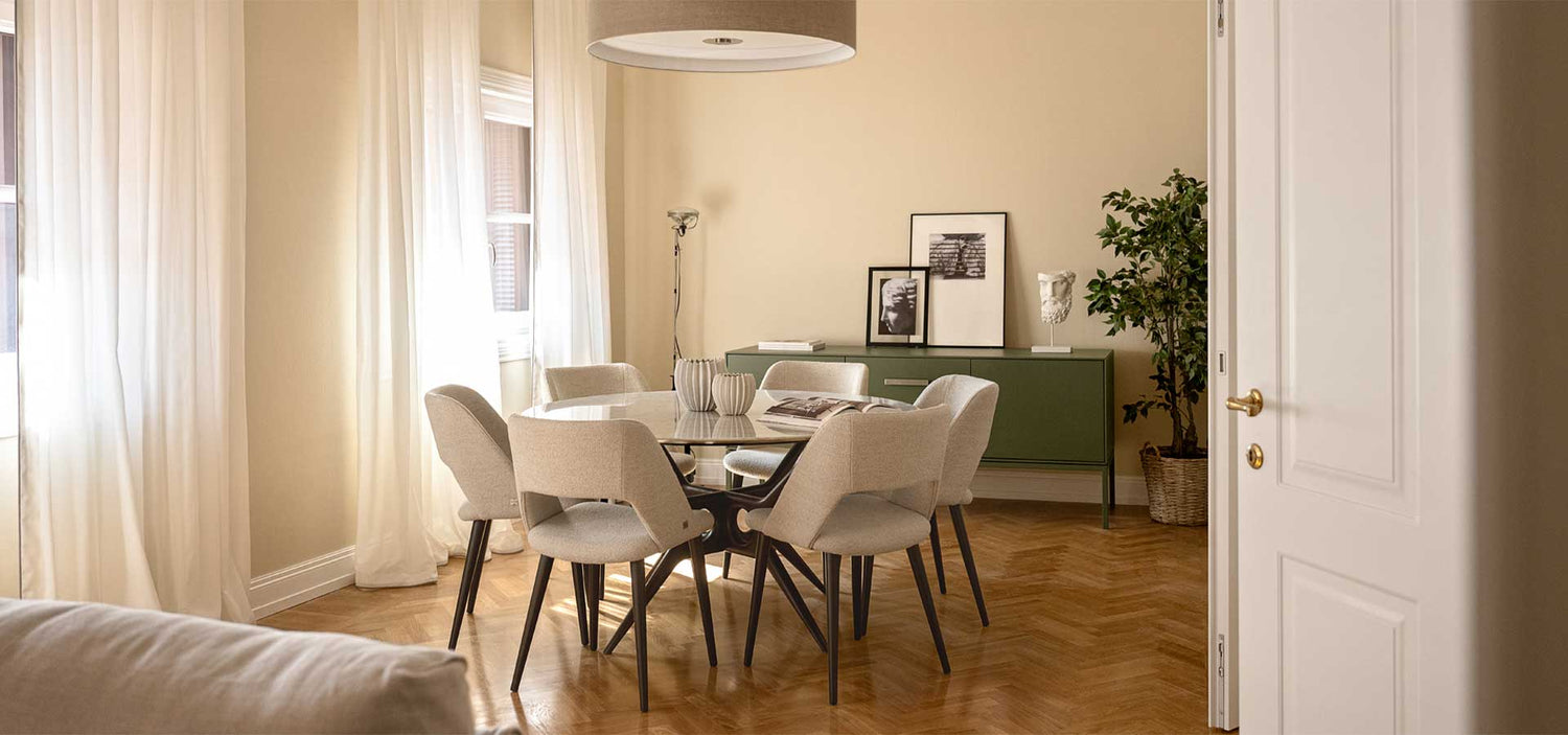 Zona pranzo moderna con tavolo rotondo in vetro, sei sedie chiare, pavimento in parquet e arredi minimal in un ambiente luminoso.